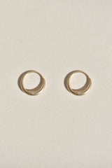 A pair of small gold huggie hoop earrings in satin finish on a textured off-white background.