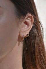 A close-up of a person's ear wearing three small gold hoop earrings, with brown hair and a light neutral background.