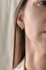 A close-up of a woman's ear wearing a small gold huggie hoop earring, with her face and hair partially visible against a soft neutral background.