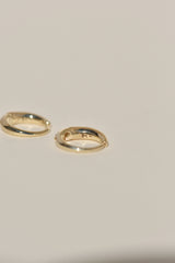 Two small gold huggie hoop earrings on a plain light beige background.